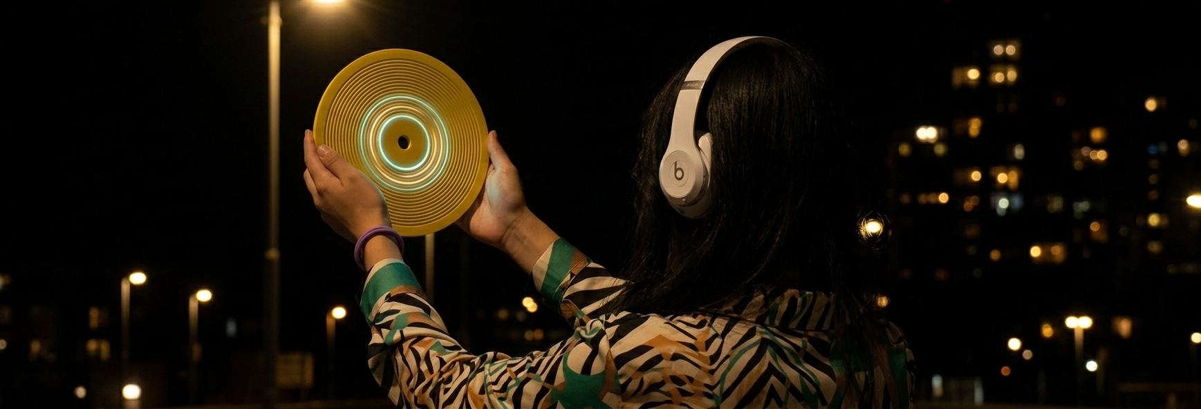 A listener wearing headphones holds a gold Kräftsound disc against the city lights at night